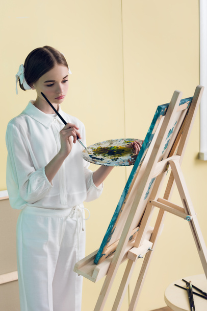 pensive teenager painting on easel with brush and palette  - Photo, Image
