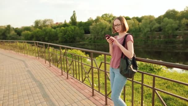 Trendikäs tyttö laseissa älypuhelimella Euroopan kaupungissa. Hän seisoo ulkona sillalla. Auringonlasku
. - Materiaali, video