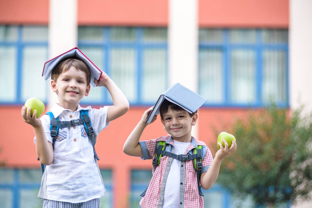 Child going to school. two boy friend holding books on head house roof, first school day. Little students excited to be back to school. Beginning of class vacation. Kids eating apple in yard - Photo, Image