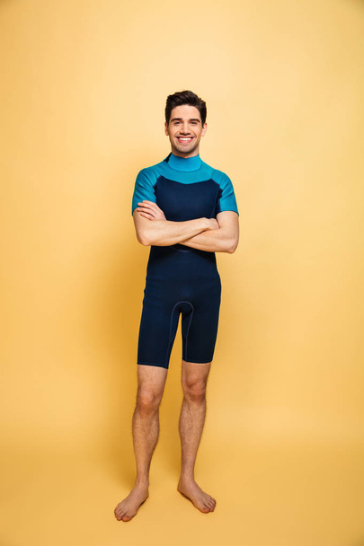 Full length portrait of a happy young man dressed in swimsuit standing with arms folded isolated over yellow background - Foto, Imagen