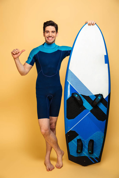 Portrait of a happy young man dressed in swimsuit holding surfboard isolated over yellow background pointing. - Фото, зображення