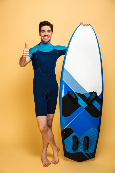 Portrait of a cheerful young man dressed in swimsuit holding surfboard isolated over yellow background make thumbs up. - Fotoğraf, Görsel
