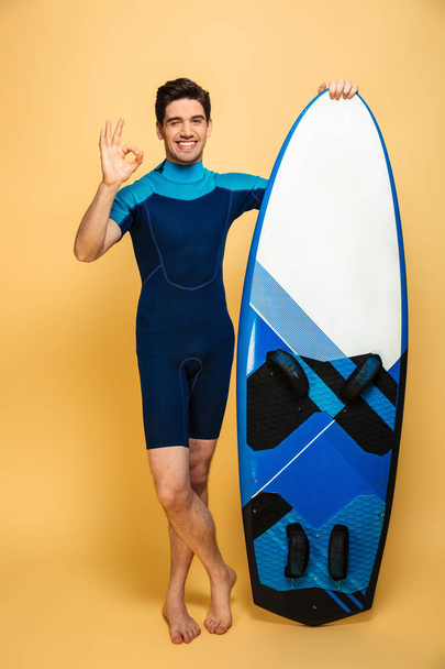 Portrait of a cheerful young man dressed in swimsuit holding surfboard isolated over yellow background make okay gesture. - Foto, afbeelding