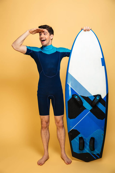 Full length portrait of a happy young man dressed in swimsuit holding surfboard and looking far away isolated over yellow background - Foto, imagen