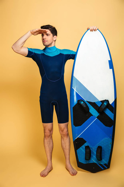 Full length portrait of an excited young man dressed in swimsuit holding surfboard and looking far away isolated over yellow background - Фото, зображення