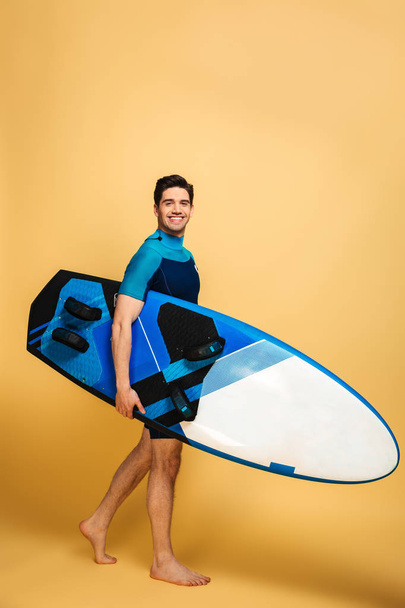 Full length portrait of a smiling young man dressed in swimsuit walking with a surfboard isolated over yellow background - Fotó, kép