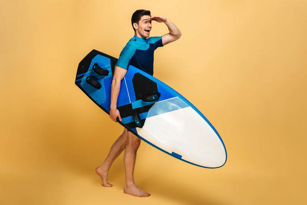 Bild eines aufgeregten jungen Mannes im Badeanzug, der sein Surfbrett isoliert vor gelbem Hintergrund zur Seite hält. - Foto, Bild
