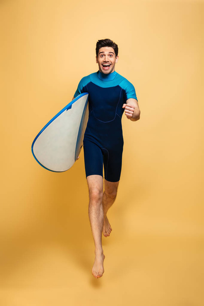 Full length portrait of a happy young man dressed in swimsuit running with a surfboard isolated over yellow background - Фото, изображение