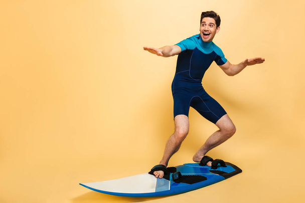 Full length portrait of a happy young man dressed in swimsuit surfing on a board isolated over yellow background - Foto, Imagen