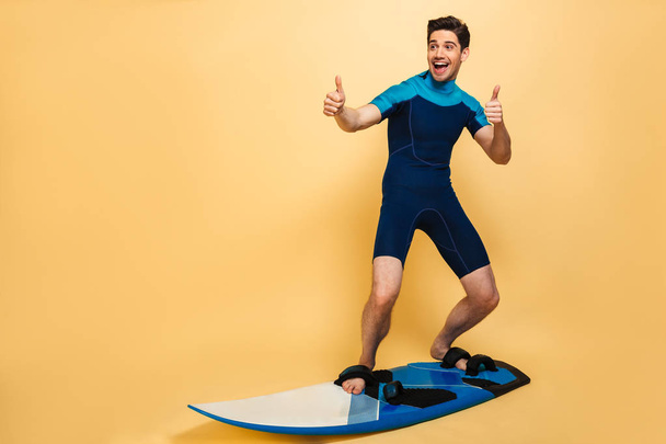 Full length portrait of a happy young man dressed in swimsuit surfing on a board and showing thumbs up isolated over yellow background - Foto, afbeelding