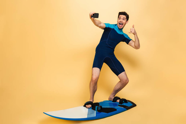 Full length portrait of a confident young man dressed in swimsuit taking a selfie while surfing on a board isolated over yellow background - Foto, immagini