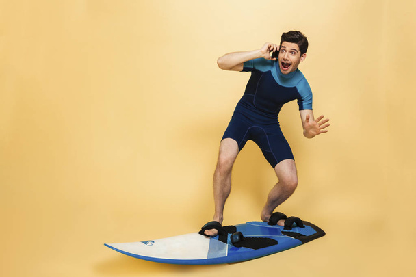 Photo of a screaming scared young man dressed in swimsuit isolated over yellow background on surfing board talking by phone. - Foto, immagini