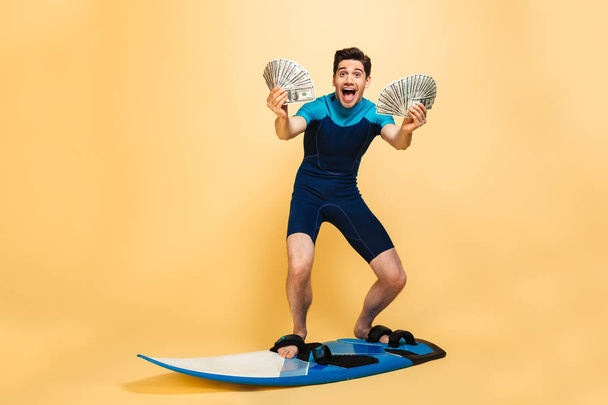 Full length portrait of an excited young man dressed in swimsuit holding bunch of money banknotes while surfing on a board isolated over yellow background - Фото, зображення