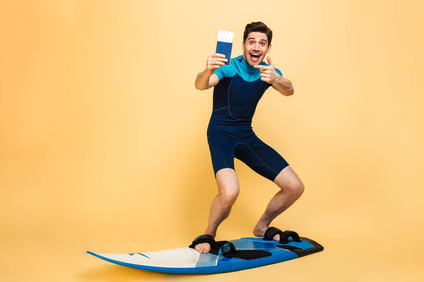 Ganzkörperporträt eines glücklichen jungen Mannes im Badeanzug, der mit dem Finger auf den Pass zeigt, während er auf einem Brett surft, unter der Gelddusche isoliert vor gelbem Hintergrund - Foto, Bild