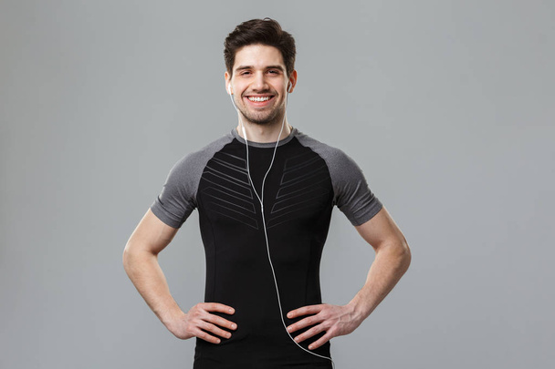 Photo of happy young sportsman standing isolated over grey wall background looking camera listening music with earphones. - Φωτογραφία, εικόνα