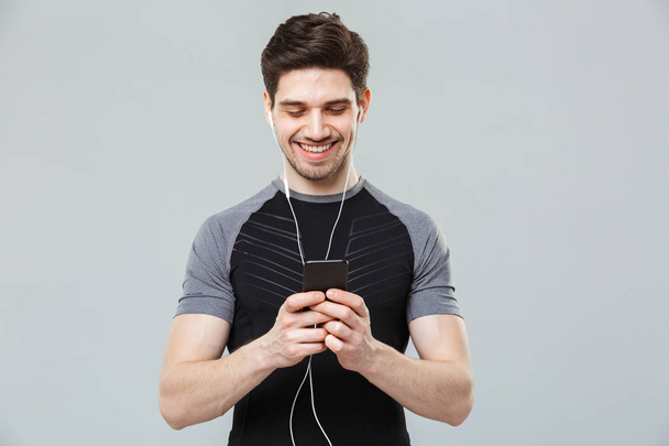 Portrait of a smiling young sportsman listening to music with earphones while using mobile phone isolated over gray background - Fotó, kép