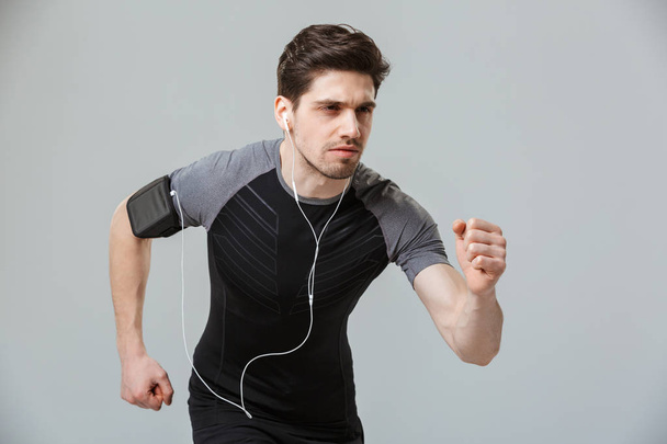 Portrait of a concentrated young sportsman listening to music with earphones while running isolated over gray background - Foto, Imagem