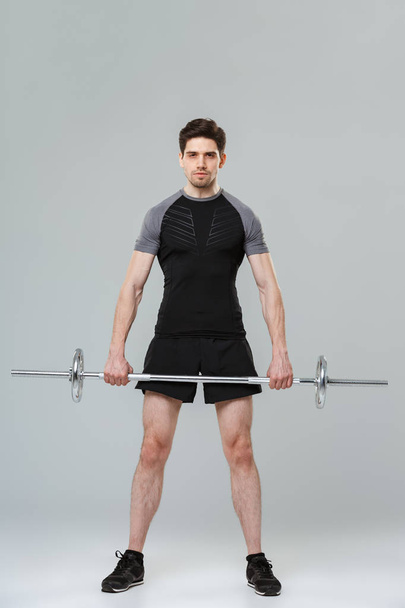 Full length portrait of a concentrated young sportsman lifting heavy barbell isolated over gray background - Фото, изображение