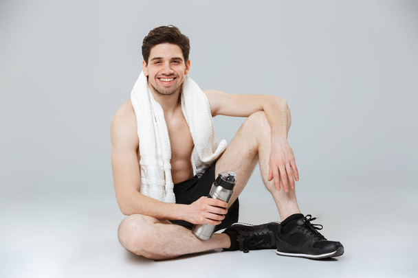 Portrait of a smiling young half naked sportsman resting while sitting with towel and water bottle isolated over gray background - Fotó, kép