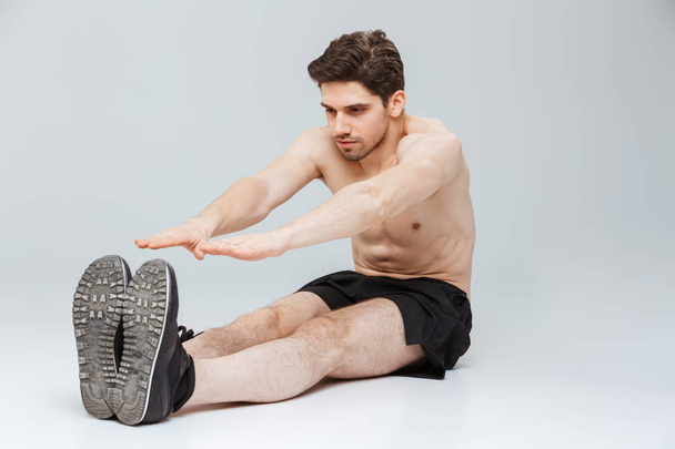 Portrait of a confident young half naked sportsman doing stretching exercises while sitting isolated over gray background - Foto, afbeelding