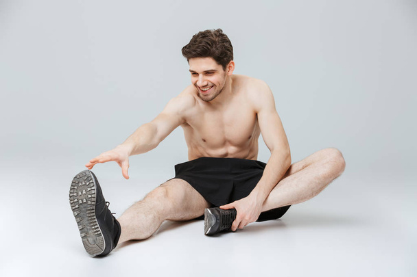 Portrait of a fit young half naked sportsman doing stretching exercises while sitting isolated over gray background - Fotoğraf, Görsel