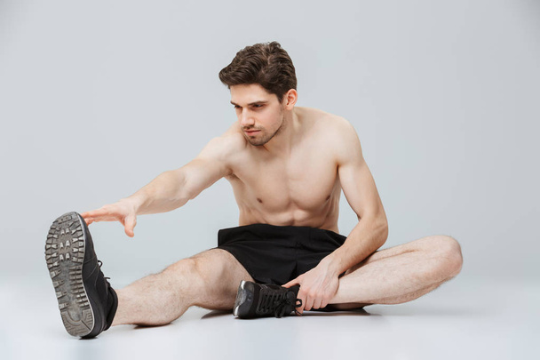 Πορτρέτο του ένα αυτοπεποίθηση νέους μισό γυμνό αθλητή που κάνει διατάσεις ενώ κάθεται απομονωμένος σε γκρι φόντο - Φωτογραφία, εικόνα