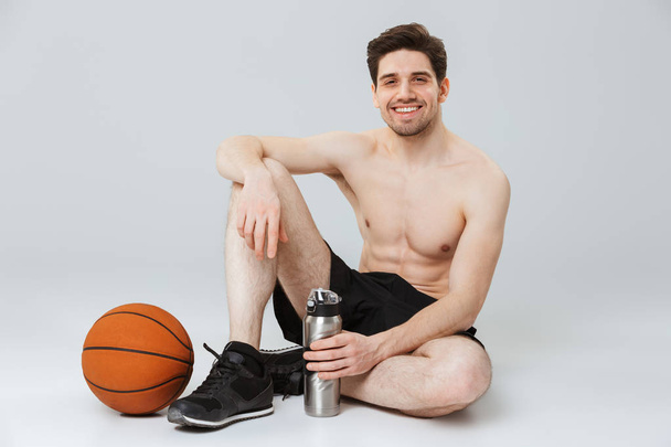 Retrato de un joven deportista semidesnudo feliz sosteniendo una botella de agua mientras está sentado con baloncesto aislado sobre fondo gris
 - Foto, Imagen
