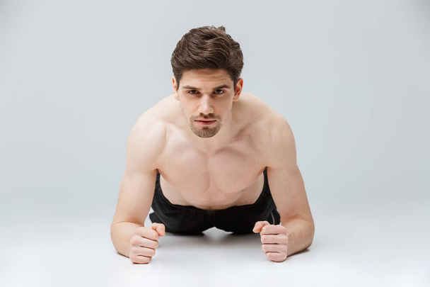 Portrait of a confident young half naked sportsman doing plank exercises over gray background - Foto, Imagem