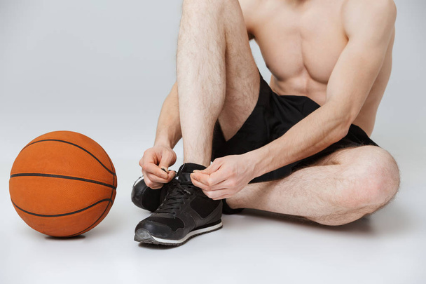 Обрезанное изображение молодого полуобнаженного спортсмена, завязывающего шнурки, сидящего с баскетбольным изолированным на сером фоне
 - Фото, изображение