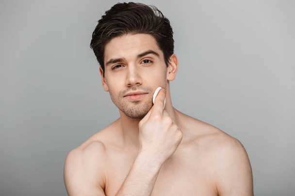 Beauty portrait of half naked handsome young man using cotton pad on his face isolated over gray background - Fotoğraf, Görsel
