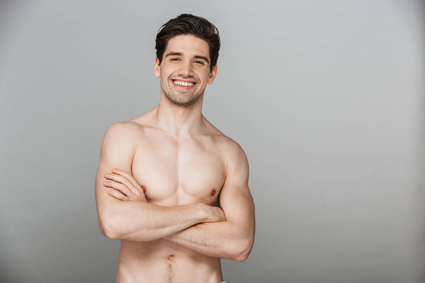 Beauty portrait of half naked smiling young man looking at camera with arms folded isolated over gray background - Foto, Imagen