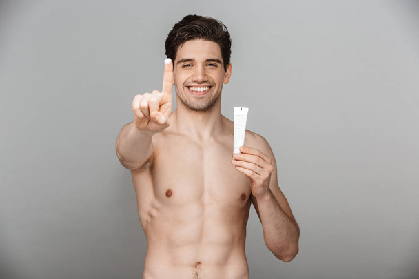 Beauty portrait of half naked confident young man showing facial cream tube isolated over gray background - Photo, image