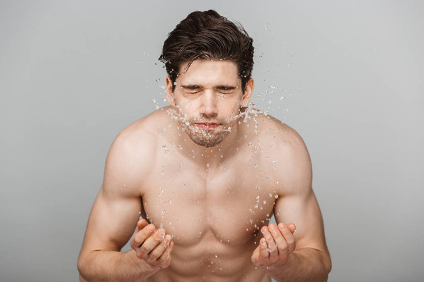 Retrato de belleza de un joven guapo medio desnudo lavándose la cara aislado sobre un fondo gris
 - Foto, Imagen