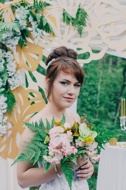schöne Frau im Hochzeitskleid auf einer mit frischen Blumen dekorierten Fotozone, posiert und porträtiert. - Foto, Bild