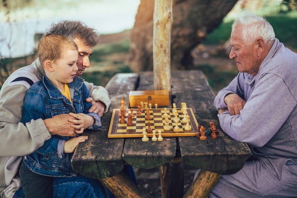 aktive Rentner, alte Freunde und Freizeit, zwei ältere Männer, die Spaß haben und im Park Schach spielen, Zeit mit dem Enkel verbringen - Foto, Bild