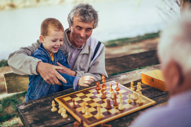 aktive Rentner, alte Freunde und Freizeit, zwei ältere Männer, die Spaß haben und im Park Schach spielen, Zeit mit dem Enkel verbringen - Foto, Bild