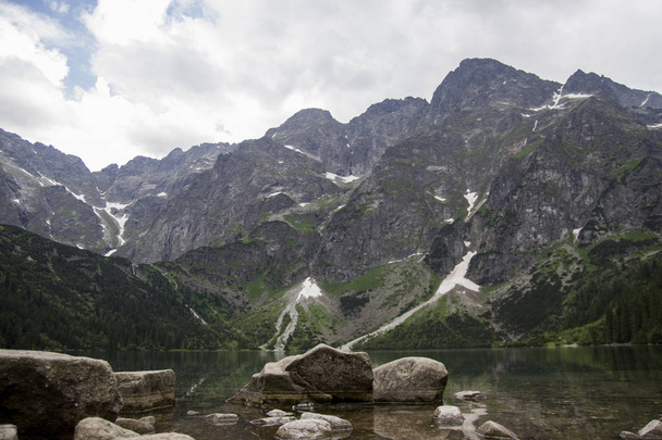 Hermosa vista del lago alpino de montaña de verano cubierto de árboles verdes con piedras en frente y nubes en el cielo. Reflejo de montaña en el agua. Agua cristalina. Europa, Alpes
 - Foto, Imagen