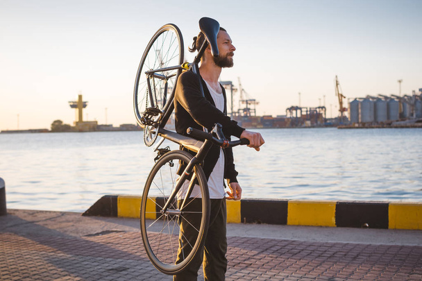 Stylish handsome man walking with bicycle on his shoulder during sunset or sunrise with sea port on background - Φωτογραφία, εικόνα