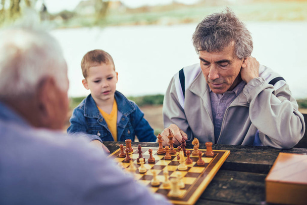 Des retraités actifs, de vieux amis et du temps libre, deux hommes âgés qui s'amusent et jouent aux échecs au parc, passent du temps avec leur petit-fils
 - Photo, image
