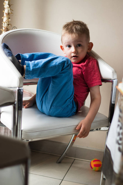 Счастливый и игривый маленький двухлетний мальчик, одетый в пижаму, сидит на белом стуле в студии фотографа, поднимая ноги и позируя. Дети и счастье концепции. Изолированный выстрел, горизонтальный
 - Фото, изображение