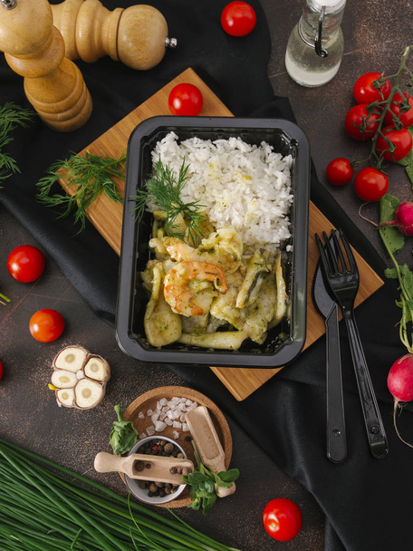 Food composition of rice with roasted zucchini and shrimps in black tray on wooden board on table with vegetables and kitchenware - Foto, imagen