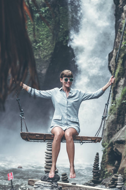 Hermosa mujer oscila cerca de la cascada en la selva de la isla de Bali, Indonesia
. - Foto, Imagen