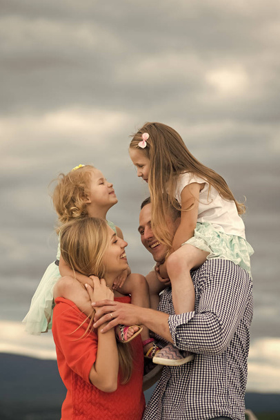 Mothers, fathers day. Children and parents enjoy vacation together. Girls sit on mother father shoulders on cloudy sky. Summer, adventure, discovery, wanderlust concept - 写真・画像