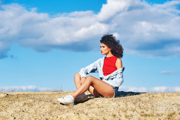 Young woman sitting on sand on sky background . Girl in shorts and shirt in nature - Photo, Image