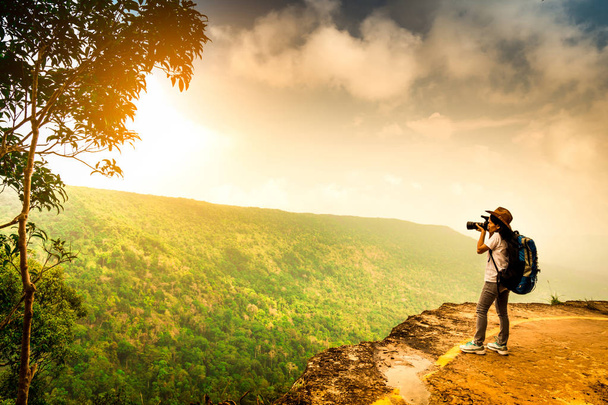 Mujer joven viajera con mochila, sombrero y cámara de pie en el acantilado de la montaña. Fotógrafa tomando fotos de bosque tropical, cielo y nubes en sus vacaciones. Mujer asiática viajando sola
. - Foto, Imagen