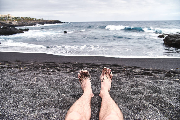 ビーチで男性の足。黒い火山砂とユニークなビーチ。波は岸のスプラッシュ、新鮮な海の風が吹いています。男、男、少年はビーチで休憩. - 写真・画像
