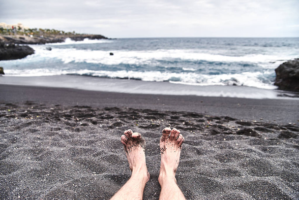 Αντρικά πόδια στην παραλία. Μια μοναδική παραλία με μαύρη ηφαιστειακή άμμο. Κύματα splash σχετικά με την ακτή, φυσά ένα φρέσκο αεράκι της θάλασσας. Ένας άνθρωπος, ένας άντρας, ένα αγόρι είναι ανάπαυση στην παραλία. - Φωτογραφία, εικόνα