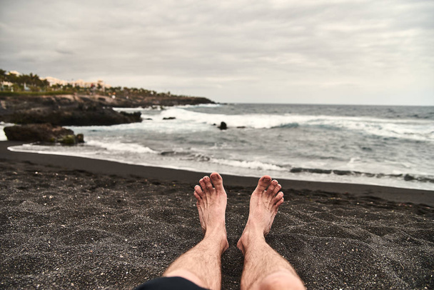 Sahilde erkek bacaklar. Siyah volkanik kum ile benzersiz bir plaj. Dalgalar kıyıya hakkında sıçrama, taze deniz meltemi esiyor. Bir adam, bir adam, bir çocuk sahilde dinleniyor. - Fotoğraf, Görsel