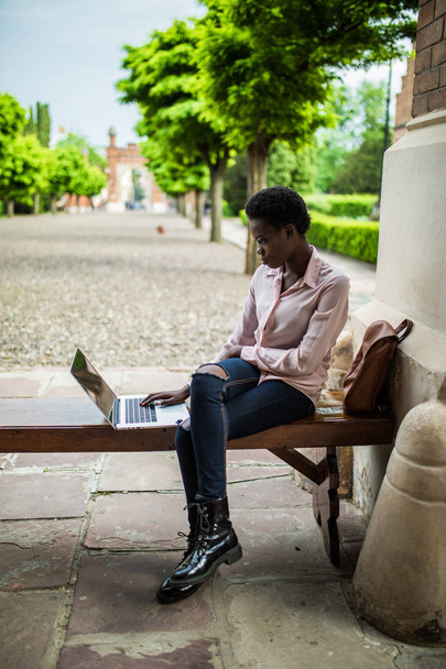 Keskittynyt nuori Afrikkalainen nainen college opiskelija etsii syvälle ajatteluun istuessaan penkillä työskentelee kannettavan kampuksella valmistautuu tentti
 - Valokuva, kuva