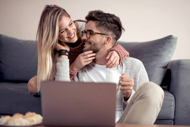 Relajada pareja joven amorosa con tazas de café utilizando el ordenador portátil en el sofá en casa
. - Foto, imagen